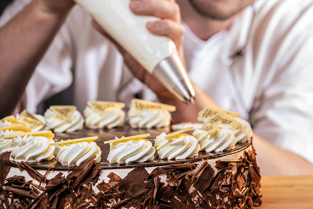 Bäcker hat einen Spritzbeutel in der hand und spritzt Sahnekringel auf eine Torte.
