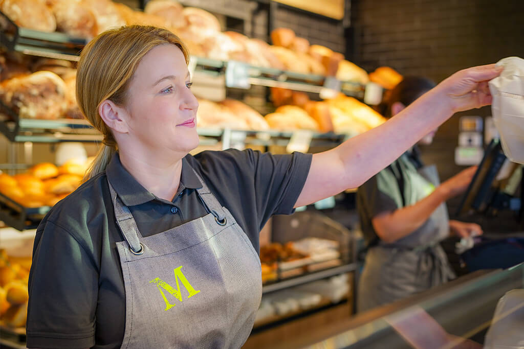 Verkäuferin steht in einer Filiale der Heidebäckerei Meyer vor einem Regal voller Brote und reicht eine Tüte mit Brötchen über die Theke.
